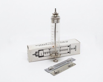 Puede incluir: Una jeringa de vidrio transparente con émbolo y punta de metal, marcada con una capacidad de 20 ml. Se coloca frente a su caja de cartón blanco original con un dibujo lineal negro de la jeringa y texto, junto a dos pequeñas agujas de metal.