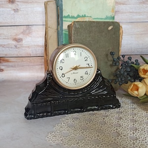 May include: A black clock with a gold face and white hands. The clock is on a black stand with a decorative design. The clock face reads "Max" and has the numbers 1 through 12.