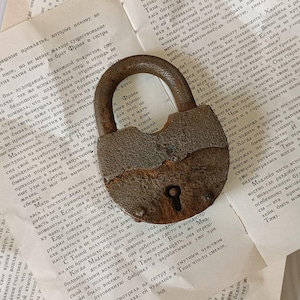 May include: A rusty metal padlock with a keyhole and a hasp. The padlock is lying on a page of a book with text in Russian.