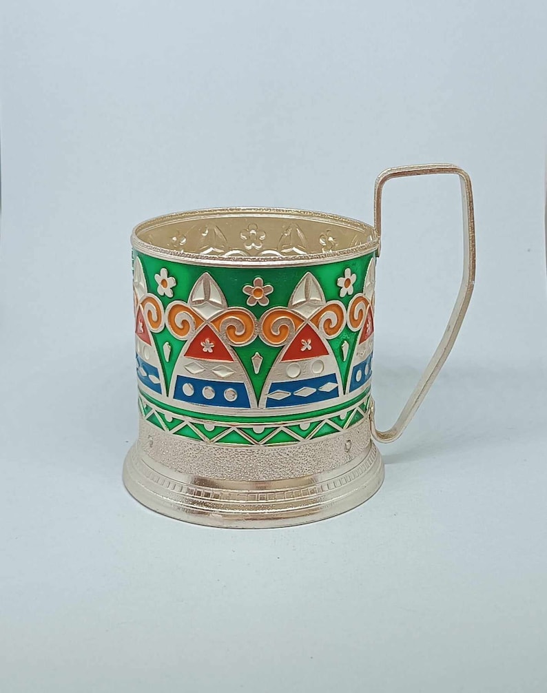 May include: Silver-plated cup holder with a green, orange, red, blue, and white geometric design. The cup holder has a handle and a textured base.