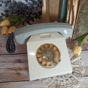 May include: A white rotary dial phone with a gray handset. The phone has a round dial with numbers 1 through 9 and 0. The phone is sitting on a wooden surface with a white doily.