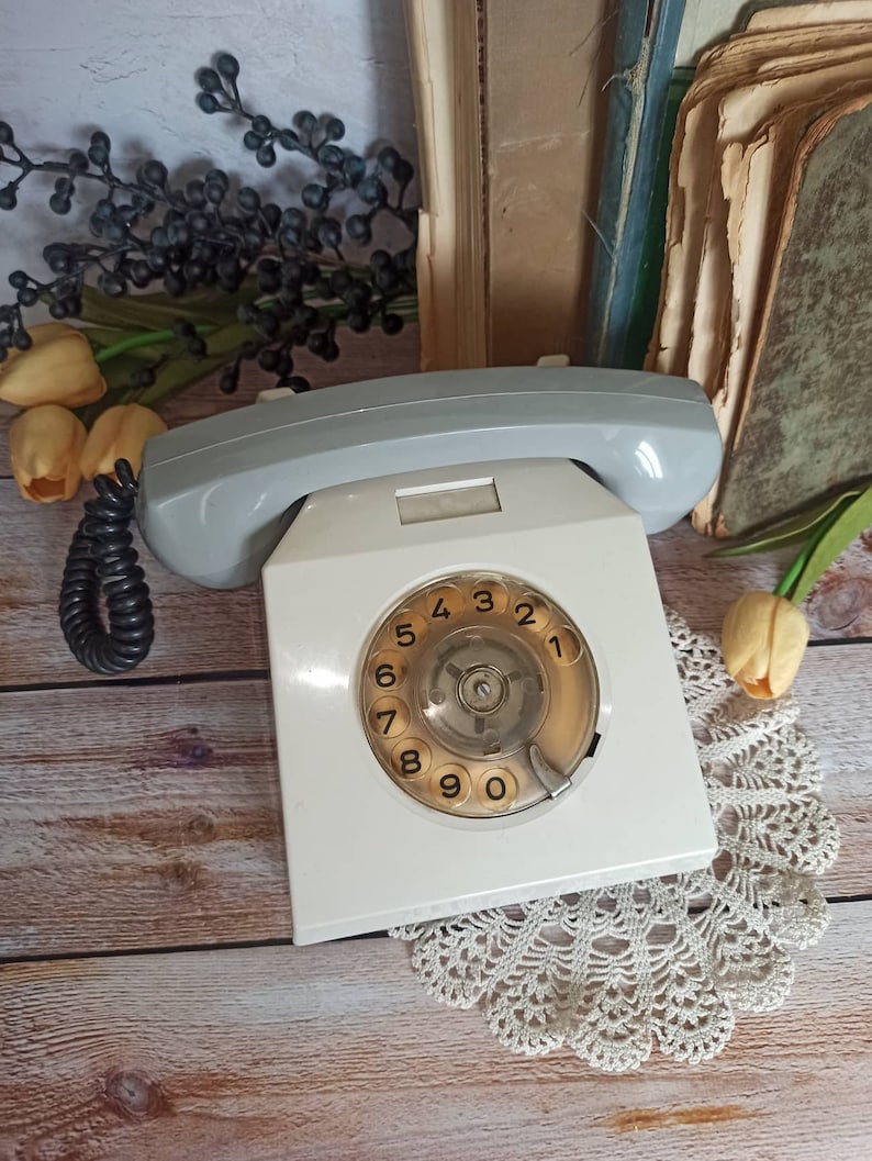 May include: A white rotary dial phone with a gray handset. The phone has a round dial with numbers 1 through 9 and 0. The phone is sitting on a wooden surface with a white doily.