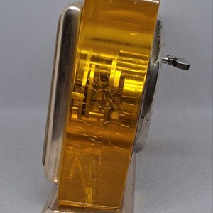 May include: Vintage Art Deco-style clock with a silver-tone metal frame and a gold-colored, transparent face. The clock has a key for winding and a rectangular base. The face shows internal mechanisms.