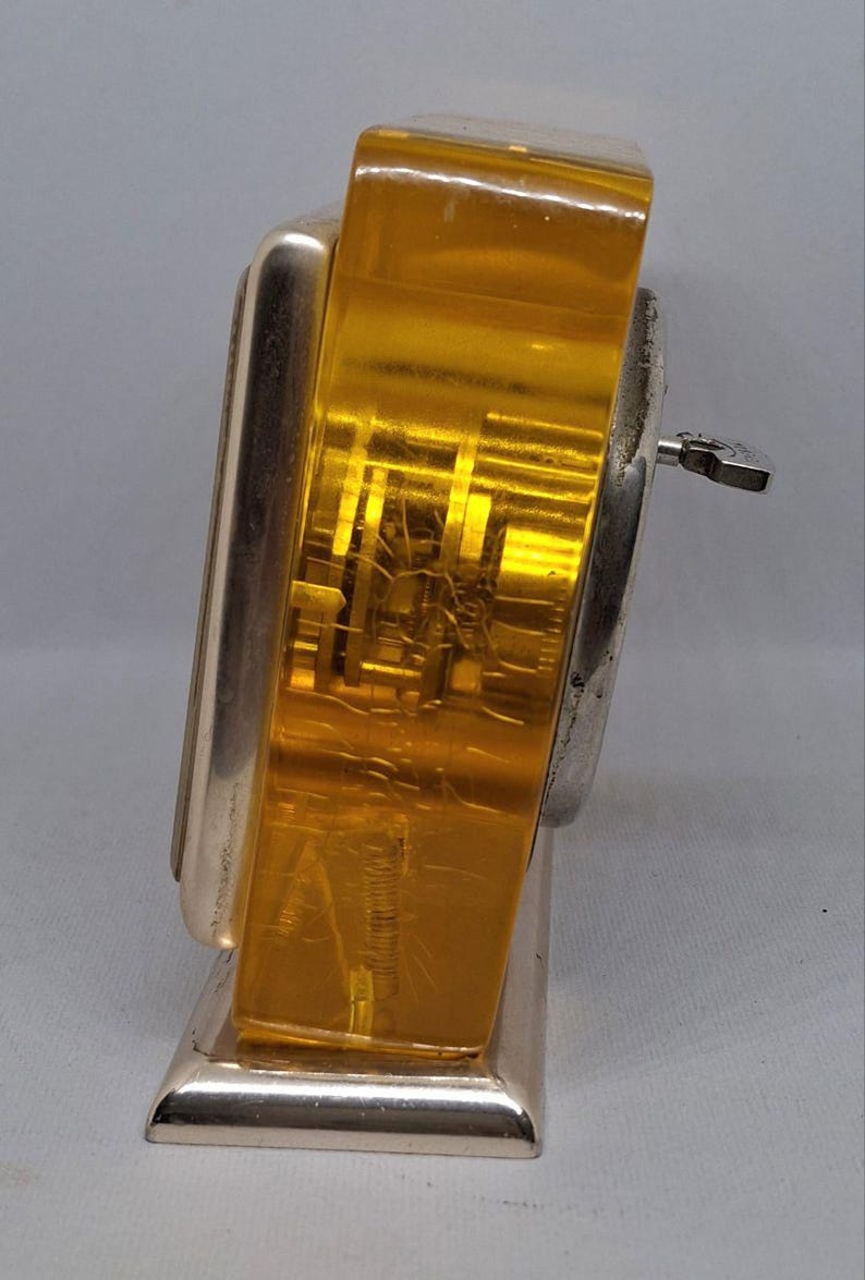 May include: Vintage Art Deco-style clock with a silver-tone metal frame and a gold-colored, transparent face. The clock has a key for winding and a rectangular base. The face shows internal mechanisms.