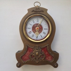Puede incluir: Un reloj de color dorado adornado con una esfera blanca y números romanos negros. El reloj tiene una base de mármol rojo y un marco dorado.