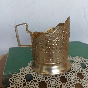 May include: A gold-colored metal cup holder with an ornate floral design. The holder has a handle and a scalloped rim.