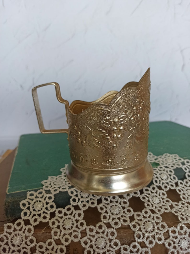 May include: A gold-colored metal cup holder with an ornate floral design. The holder has a handle and a scalloped rim.