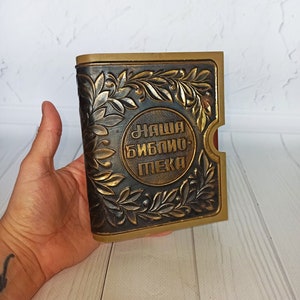 Op de afbeelding: Een klein, donkerbruin boek met gouden reliëfletters met de tekst "Наша Библию Пека". Het boek is versierd met een gouden bloemmotief.