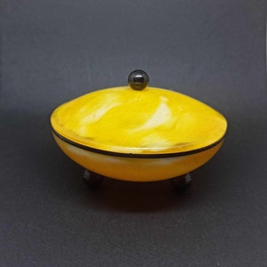 May include: A yellow and white swirl design lidded dish with a black knob on top and three black feet. The dish is made of ceramic and has a glossy finish.