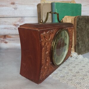 May include: A vintage wooden clock with a carved design and a brass handle. The clock face has Roman numerals and a brass frame.