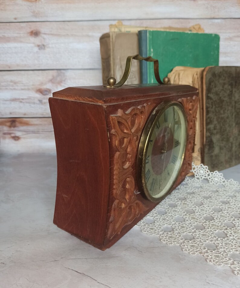 May include: A vintage wooden clock with a carved design and a brass handle. The clock face has Roman numerals and a brass frame.