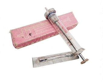 Puede incluir: Una jeringa de vidrio vintage con émbolo y punta de metal, que presenta marcas de medición negras de hasta 5 ml. Se acompaña de una caja de cartón rosa rectangular con dibujos lineales impresos y una pequeña pieza accesoria de metal plateado.