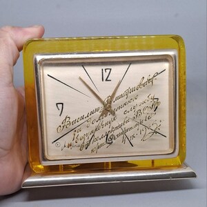 May include: A vintage rectangular clock with a silver frame and a yellow transparent border. The clock face is off-white with black numerals and hands. Handwritten text is visible on the clock face. The clock is held by a hand.