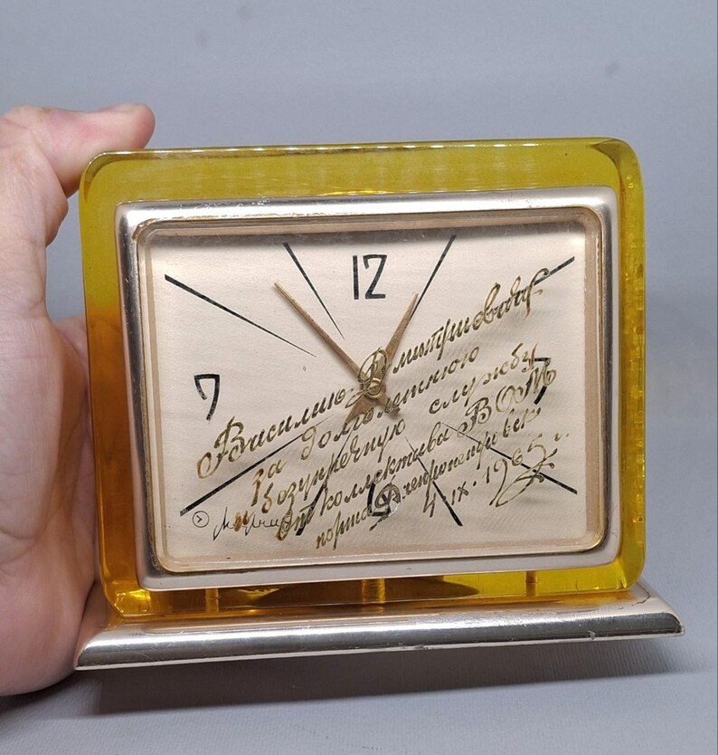 May include: A vintage rectangular clock with a silver frame and a yellow transparent border. The clock face is off-white with black numerals and hands. Handwritten text is visible on the clock face. The clock is held by a hand.