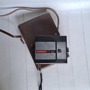 以下が含まれることがあります： 茶色の革製のケースとストラップが付いた、黒と銀の8mmフィルムカメラ。カメラには「АВРОРА 10」という文字が印刷されています。