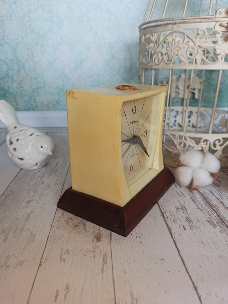 May include: A vintage cream-colored alarm clock with a brown base. The clock face has black hands and black numbers. The clock says "Sevan" on the face.