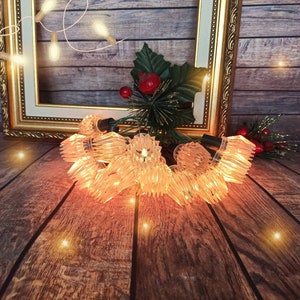 May include: A string of clear, crystal-like Christmas lights with warm white bulbs. The lights are arranged in a circular pattern and are illuminated, casting a warm glow.