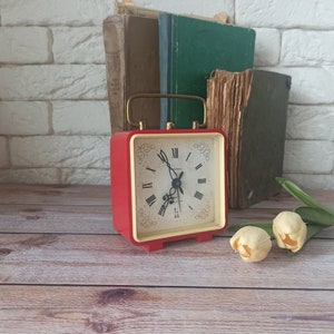 May include: A red square alarm clock with a gold handle and a cream-colored face featuring black Roman numerals. The clock is set on a wooden surface, with old books in the background and two cream-colored tulips.