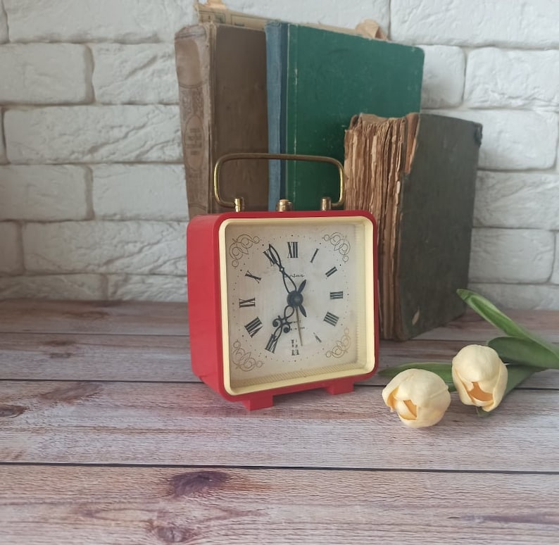May include: A red square alarm clock with a gold handle and a cream-colored face featuring black Roman numerals. The clock is set on a wooden surface, with old books in the background and two cream-colored tulips.