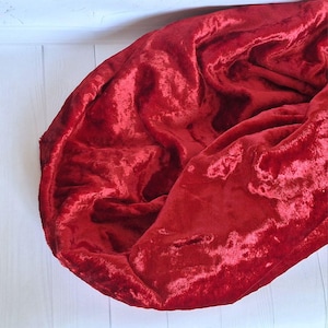 May include: A luxurious, deep red velvet fabric is draped, showcasing its rich texture and sheen. The fabric is folded, creating shadows and highlights that emphasize its plushness. The background is a light, wooden surface.