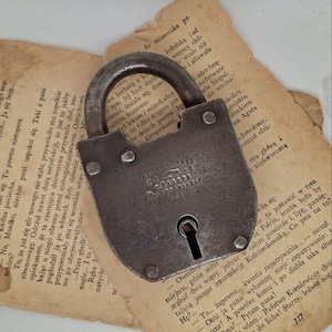 May include: A worn metal padlock with a curved handle, on top of aged, yellowed book pages. The lock has a keyhole and is riveted. The background features Polish text.