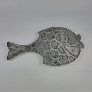 May include: A silver-colored, fish-shaped trivet with a detailed scale pattern. The trivet has a tail fin and a smiling face, adding a decorative touch to kitchenware. The metal trivet is designed to protect surfaces from hot cookware.