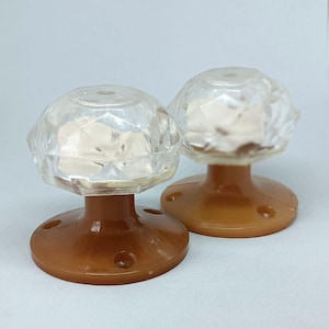 Może przedstawiać: Dwa zabytkowe, przezroczyste, fasetowane szklane gałki do drzwi z brązowymi podstawami. Gałki mają wzór przypominający diament i są zamontowane na okrągłych, brązowych podstawach z trzema otworami na śruby. Gałki są prawdopodobnie do szafki lub szuflady.