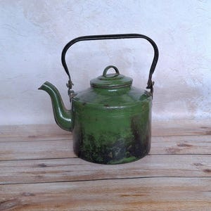 Könnte beinhalten: Vintage grüne Emaille-Teekanne mit schwarzem Griff und Ausguss. Die Teekanne hat einen abgerundeten Körper und einen Deckel mit einem kleinen Schlaufengriff. Die Farbe weist Gebrauchsspuren auf. Ein klassischer Küchenartikel.