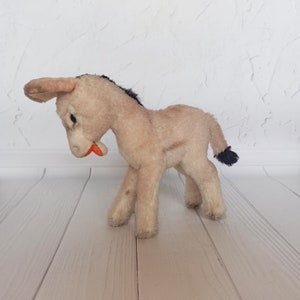 May include: A plush donkey toy with a light brown body, black mane and tail, and an orange mouth. The donkey is standing on a light wood floor.