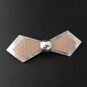 May include: A silver bow tie brooch with a beige center featuring a geometric pattern. The center of the bow is accented with a silver ball.