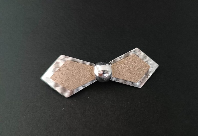 May include: A silver bow tie brooch with a beige center featuring a geometric pattern. The center of the bow is accented with a silver ball.