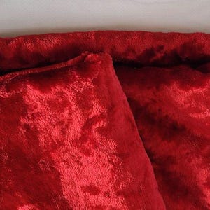 May include: Close-up of a folded piece of red velvet fabric. The plush material has a soft, textured surface with a slight sheen. The fabric is folded over itself, revealing the texture and color.