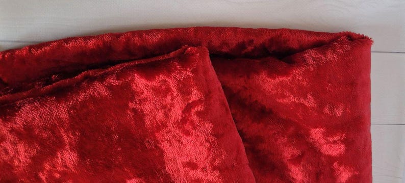 May include: Close-up of a folded piece of red velvet fabric. The plush material has a soft, textured surface with a slight sheen. The fabric is folded over itself, revealing the texture and color.