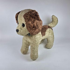 Op de afbeelding: Een vintage pluchen hondenspeelgoed met een beige lichaam en bruine oren en vlekken. De hond heeft zwarte ogen en een kleine blauwe kraag. Het pluche speelgoed staat op alle vier de poten met zijn staart omhoog.