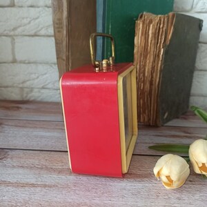 May include: A vintage red and cream-colored alarm clock with a gold handle. The clock has a rectangular shape and sits on a wooden surface. Old books are in the background.