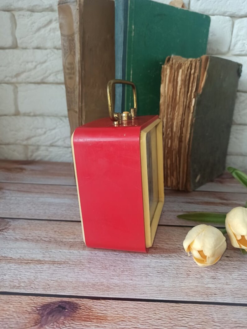 May include: A vintage red and cream-colored alarm clock with a gold handle. The clock has a rectangular shape and sits on a wooden surface. Old books are in the background.