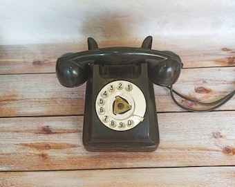 May include: An antique black rotary dial telephone with a white dial and black handset. The phone sits on a wooden surface. The dial features numbers 0-9. The phone has a classic, vintage design.