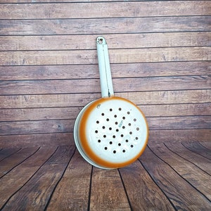Può includere: Scolapasta in smalto bianco con bordo arancione e manico. Lo scolapasta ha una forma rotonda e un motivo di fori. Lo scolapasta è appoggiato su una superficie di legno.