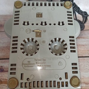May include: The bottom of a gray rotary phone with a black cord. The phone has a label that reads "Variant Typ: 116-10551", "Baujahr: 6/71", "Fernmeldewerk Nordhausen", "Made in Germany".