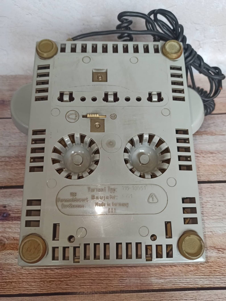 May include: The bottom of a gray rotary phone with a black cord. The phone has a label that reads "Variant Typ: 116-10551", "Baujahr: 6/71", "Fernmeldewerk Nordhausen", "Made in Germany".