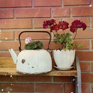 以下が含まれることがあります： 白いエナメル製のティーポットの中にピンクの花が1つ、白い陶器製の鉢の中にピンクの花がいくつか入っています。どちらもレンガの壁に面した木の棚の上に置かれています。