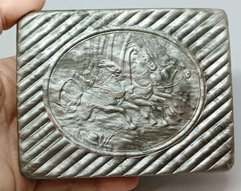 May include: A silver-colored, rectangular box with a raised, circular design featuring a scene of three horses pulling a carriage through a forest. The box has a textured, diagonal pattern.