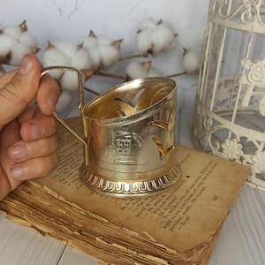 May include: A vintage gold-colored cup holder with a handle, featuring cut-out bird designs and a detailed building illustration. The cup holder rests on an aged book with visible text, suggesting a historical theme.