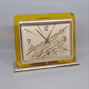 May include: A vintage desk clock with a silver frame and a yellow acrylic border. The clock face is cream-colored with black numerals and hands. Handwritten script covers the face. The clock sits on a silver base.