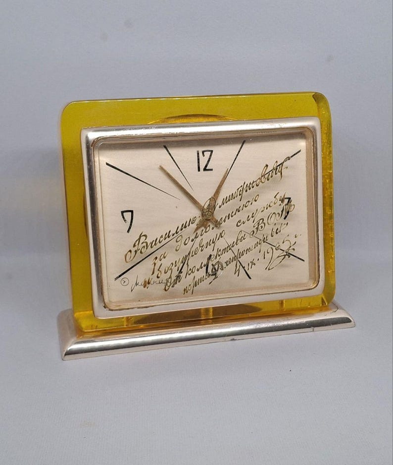 May include: A vintage desk clock with a silver frame and a yellow acrylic border. The clock face is cream-colored with black numerals and hands. Handwritten script covers the face. The clock sits on a silver base.