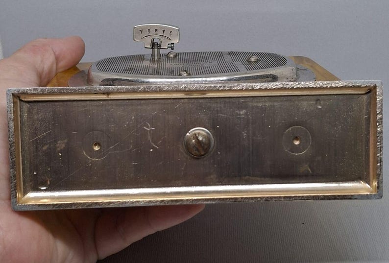 May include: A vintage silver-toned music box with a turntable design. The box has a rectangular base with a silver frame and a wooden top. The turntable has a silver arm and a small handle. The word "TORVC" is visible on the handle.