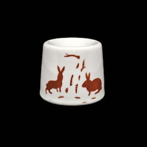 May include: A white ceramic egg cup with a brown rabbit design. The egg cup features two rabbits and stylized grass. The design is repeated around the cup. The egg cup is cylindrical with a slightly flared base.