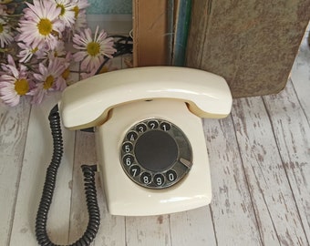 Pode incluir: Um telefone de disco branco com um dial preto e um cabo preto. O telefone está sobre uma superfície de madeira branca.