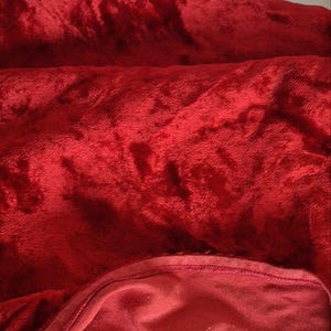 May include: A close-up of a plush, red velvet fabric. The fabric is folded and draped, showing a soft, textured surface. The color is a deep, rich red, with subtle variations in tone due to the folds and shadows. The edges of the fabric are visible.