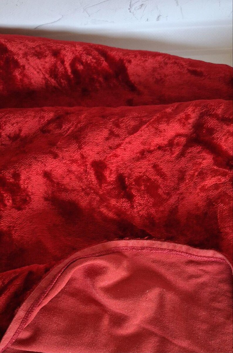 May include: A close-up of a plush, red velvet fabric. The fabric is folded and draped, showing a soft, textured surface. The color is a deep, rich red, with subtle variations in tone due to the folds and shadows. The edges of the fabric are visible.